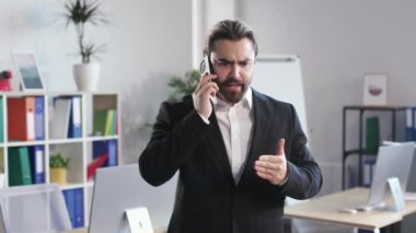 Outraged businessman talking on mobile phone with partner at modern office room. Bearded male in suit arguing and actively gesticulating during serious conversation.