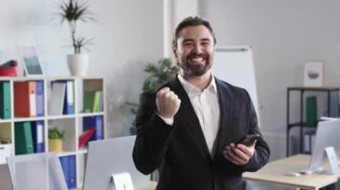 Overjoyed young man in suit holding smartphone and screaming yes with clenched fist at modern office. Bearded male reading great news and smiling while looking at camera.