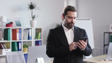 Portrait of male businessman using mobile phone for scrolling social media during work at modern office. Bearded man enjoying reading news and looking at camera with smile.