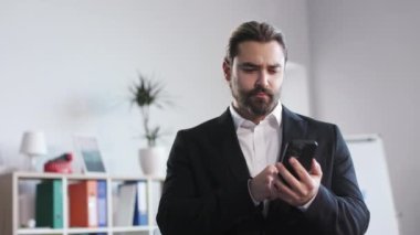 Young male office worker browsing web on smartphone during break while standing at bright company meeting room. Concept of resting with modern electronic devices.