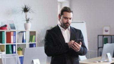Concentrated male manager using mobile phone while working at bright office. Young man in formal suit browsing internet for searching necessary information.