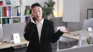 Young asian female having phone conversation while standing at modern open space office. Attractive woman in formal suit negotiating with new business client.