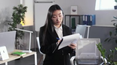 Focused asian woman in formal wear and eyeglasses standing at open space office and reading documents on clipboard. Female manager preparing speech for financial presentation on meeting.