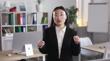 Confident female manager in stylish black suit adjusting eyewear while standing at office and looking at camera. Portrait of asian woman with dark long hair indoors.