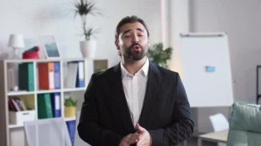 Confident bearded entrepreneur talking about company while standing at office and looking at camera. Competent business trainer recording webinar, blog and online tutorial indoors.