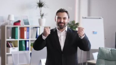 Portrait of handsome caucasian man in black suit gesturing from happiness while standing at office and looking at camera. Bearded businessman celebrating success at work.