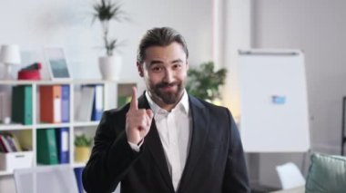 Confident smiling businessman in black suit pointing with forefinger on camera while standing open space office. Concept of people, gesture and recommendation.