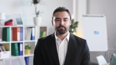 Portrait of caucasian bearded man with calm face posing at modern office. Handsome male employee dressed in stylish business suit standing at workplace.
