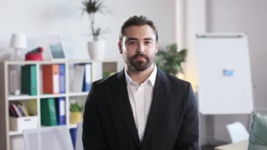 Caucasian male entrepreneur looking at camera with confidence in eyes while standing at business center room. Handsome bearded man wearing stylish black formal suit.