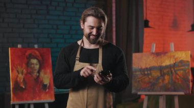 Caucasian art master holding modern smartphone and scrolling news in social network. Handsome male painter taking break from creative process with cell phone in hands.