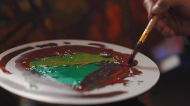 Close up of male painter mixing with brush various paints on color palette. Creative art worker making unique color for painting on canvas art workshop.