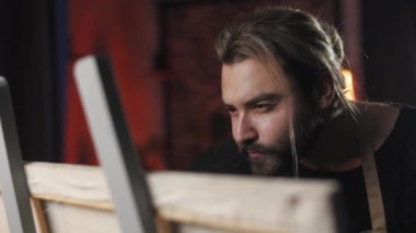 Focused bearded man standing near easel and drawing with brush and color palette. Caucasian male painter in casual attire spending free time with creativity at workshop.