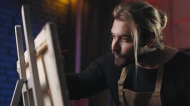 Handsome man with stylish beard and haircut using paintbrush and color palette for drawing on canvas. Male painter standing in front of easel with focused facial expression.