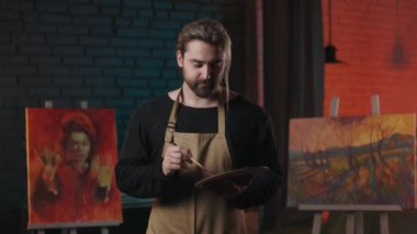 Front view of talented caucasian man in apron mixing with brush paints on wooden color palette. Bearded male painter standing in art studio, smiling and looking at camera.