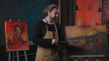 Happy bearded man standing near beautiful drawing and showing thumb up gesture. Caucasian art master in apron satisfied and proud of his masterpiece.