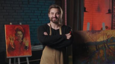 Smiling bearded male painter keeping arms crossed while standing among his masterpieces at workshop. Portrait of inspired caucasian artist.
