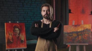 Confident male artist with paintbrush in hands standing in modern art space and looking at camera with calm face. Concept of people, creativity and inspiration.