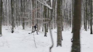 Active young guy dressed in black sport clothes spending morning time for jogging outdoors. Caucasian sporty man enjoying leisure activity among forest with snow lying around.