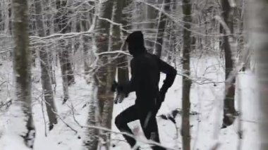 Side view of caucasian young sportsman having morning run among forest with snowy trees around. Active man wearing warm sport clothes during exercises outdoors.
