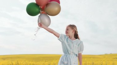 Elinde renkli hava balonlarıyla kırsal kesimde poz veren şık yaz elbiseli sevimli beyaz kız. Kaygısız dişi çocuk açık mavi gökyüzü ile vahşi sarı tarlaların arasında oynuyor..