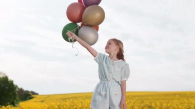 Doğal sarı saçlı güzel bir kız elinde renkli hava balonları tutarken sarı çiçeklerle yaz otlağının ortasında dikiliyor. Çocukluk kavramı ve neşeli ruh hali.