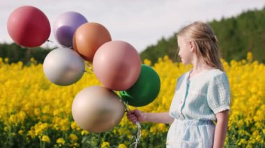 Açık havada duran, mavi gökyüzü ve arka planda sarı tarlası olan, doğal sarı saçlı, beyaz bir kız. Elinde renkli hava balonları tutan şık elbiseli, gülümseyen çocuk..