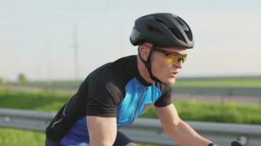Kasklı ve güneş gözlüklü formda sporcu dışarıda bisiklet sürüyor. Kırsal yolda profesyonel bisiklet süren ciddi bir erkek bisikletçi..