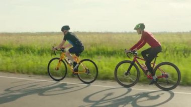 Güçlü bacakları olan kaslı adamlar kırsal kesimde profesyonel bisikletlerle uzun mesafeli yolculuk yapıyorlar. Yeşil yaz tarlası arka planda. Spor kıyafetleri ve kask giyen iki arkadaş..