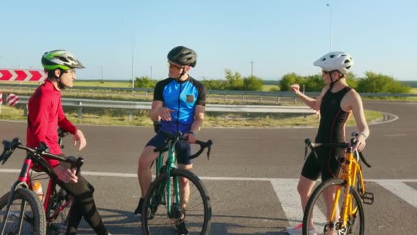 アクティブな姿の白人女性と2人の男性 彼らの自転車とチャットとアスファルトの道路に立ってスポーツヘルメットと眼鏡 新鮮な空気の中でワークアウト中に休憩を取るプロのサイクリスト — ストック動画