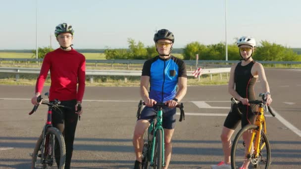 アスファルトの道路やカメラを見て上の彼らのスポーツバイクに立って2人の男性と1人の女性の肖像画 定期的な訓練と積極的な趣味 — ストック動画
