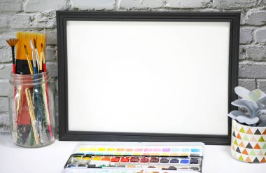 Küçük bir ev bitkisi ve suluboya malzemelerinin yanında siyah manzara çerçevesi, eskimiş beyaz tuğla duvarın önünde resim veya sanat eserlerinin bir modelini sergiliyor.