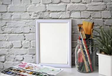 Beyaz manzara çerçevesi, küçük bir ev bitkisi ve suluboya malzemeleri, resim veya sanat eserleri, klasik beyaz tuğla duvarın önünde sergileniyor..