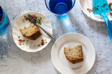 İki dilim muzlu kek beyaz tabakta çatallı ve mavi bardakta su beton masanın üstünde en üstte..