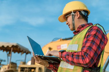 Mühendis güvenlik üniforması giyip sivil iş yapıyor. İnşaat mühendisi, kazıcıyı çalıştırmak için telsiz kullanıyor. Asyalı teknisyen bugün iş için teknoloji kullanıyor..