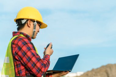 Mühendis güvenlik üniforması giyip sivil iş yapıyor. İnşaat mühendisi, kazıcıyı çalıştırmak için telsiz kullanıyor. Asyalı teknisyen bugün iş için teknoloji kullanıyor..