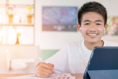 Genç Asyalı adam internette laptop ve tablet kullanıyor. Genç çocuk not defteri üzerine ders yazıyor. Ses dinleme kursu. Yeni normal Covid-19 koronavirüs. Çevrimiçi sosyal mesafe. İnternette mutlu öğrenmeler.