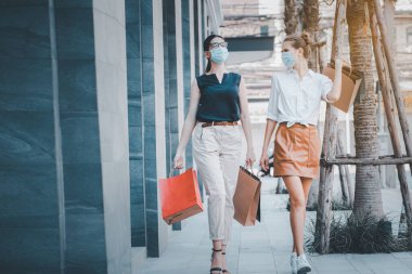 Sosyal uzaklık yeni normal kavram. Tıbbi maske takan iki mutlu kızın portresi, akşamları alışveriş merkezinin yakınında dolaşmak, alışveriş torbaları taşımak..