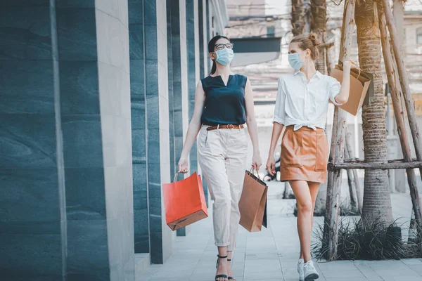 Sosyal uzaklık yeni normal kavram. Tıbbi maske takan iki mutlu kızın portresi, akşamları alışveriş merkezinin yakınında dolaşmak, alışveriş torbaları taşımak..