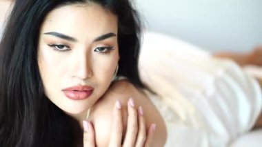 Güzel ve Seksi Asyalı Kadın Modeli 4k yatağında beyaz elbiseli kameraya poz veriyor.
