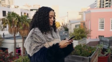 Şehirdeki bir çatıda akıllı telefondan mesajlaşan melez bir kadın. Renkli şehir manzarası ve arka planda palmiye ağaçları
