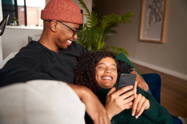 Cep telefonuna bakarken gülümseyen çok ırklı bir çift. Kanepede modern daire.