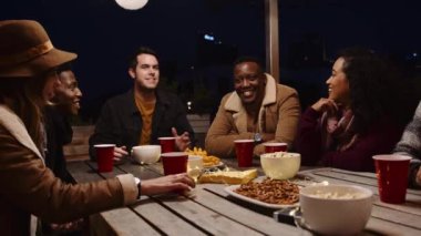 Siyahi erkek ve çeşitli yetişkin arkadaş grupları gece vakti dışarıda bir masada sohbet edip gülüyorlar.