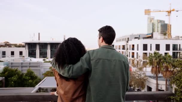 Maschio caucasico con braccio sopra la razza mista femminile in chat, guardando la vista da un tetto della città al tramonto — Video Stock