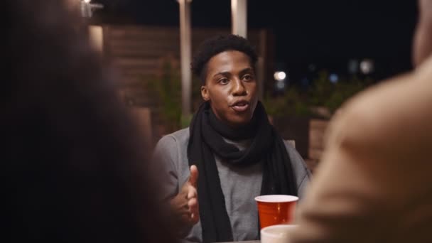 Noir adulte mâle assis à une table parler à des amis à une fête sur le toit dans la nuit. — Video
