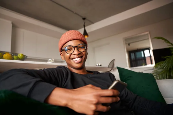 Homme noir adulte souriant à la caméra, téléphone portable à la main. Appartement moderne sur le canapé — Photo