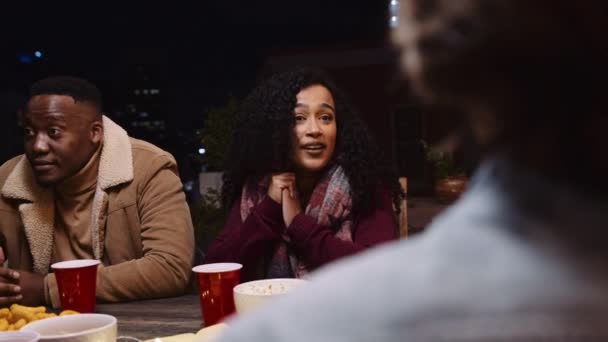 Mujer multiétnica charlando y sentada en una mesa en una fiesta en la azotea de la ciudad durante la noche — Vídeos de Stock