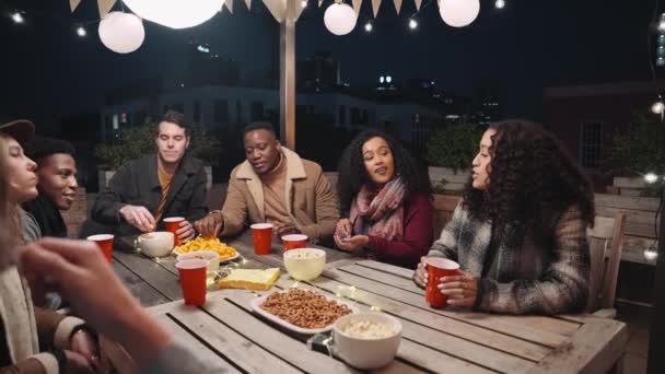 Gruppo multietnico di amici che socializzano attorno a un tavolo ad una festa sul tetto della città — Video Stock