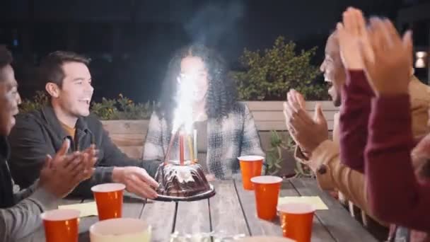 Vielfältige Gruppe von Freunden, die multiethnischen Frauen zum Geburtstag gratulieren. Wunderkerzen auf Kuchen. Nachts um einen Tisch auf einem Dach sitzen. — Stockvideo