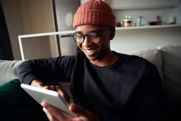 Homem negro adulto feliz sorrindo enquanto olha para tablet. Apartamento moderno no sofá — Fotografia de Stock