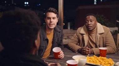 Çeşitli arkadaş grupları gece vakti çatıdaki bir partide masanın etrafında neşeli bir şekilde tartışırlar.